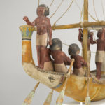 A wooden model of a boat with a crew of human figures and a passenger dressed in white clothing. Close up on figures standing at the front of the boat.