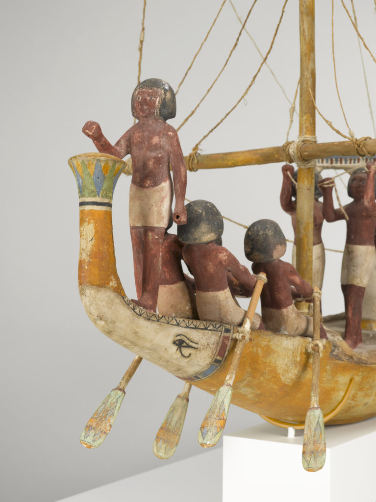 A wooden model of a boat with a crew of human figures and a passenger dressed in white clothing.