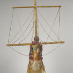 A wooden model of a boat with a crew of human figures and a passenger dressed in white clothing. Alternate view.