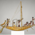 A wooden model of a boat with a crew of human figures and a passenger dressed in white clothing.
