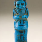 A small blue sculpture of a male figure wearing a long garment. The details of his face, wig, and hands are painted in black. There are hieroglyphs on the figure’s lower front body.