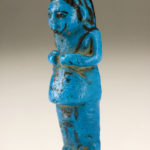 A small blue sculpture of a male figure wearing a long garment. The details of his face, wig, and hands are painted in black. There are hieroglyphs on the figure’s lower front body. Side view.