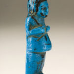 A small blue sculpture of a male figure wearing a long garment. The details of his face, wig, and hands are painted in black. There are hieroglyphs on the figure’s lower front body. Side view.