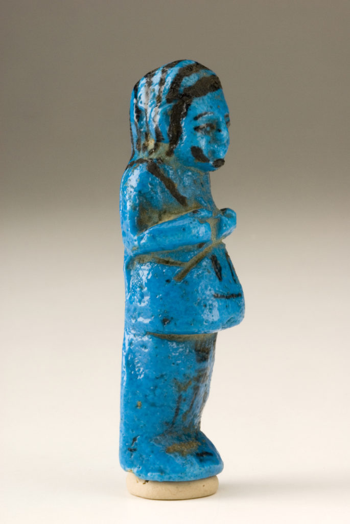 A small blue sculpture of a male figure wearing a long garment. The details of his face, wig, and hands are painted in black. There are hieroglyphs on the figure’s lower front body.