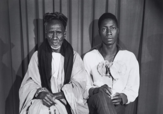 A black-and-white photograph of two Black men (possibly a father and son) sitting side by side. Their legs are crossed, and they are looking directly at the viewer. The older man is wearing a tunic and a traditional headdress. The younger man is wearing modern, slim-fitting pants and a loose, collared shirt.