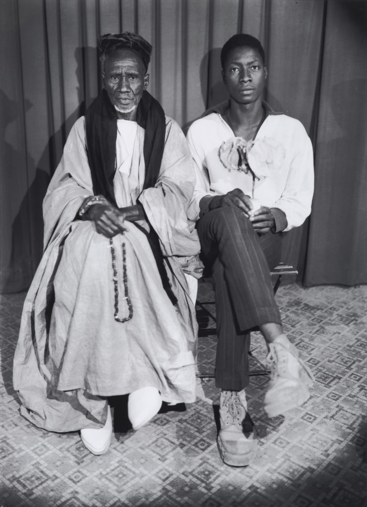 A black-and-white photograph of two Black men (possibly a father and son) sitting side by side. Their legs are crossed, and they are looking directly at the viewer. The older man is wearing a tunic and a traditional headdress. The younger man is wearing modern, slim-fitting pants and a loose, collared shirt.