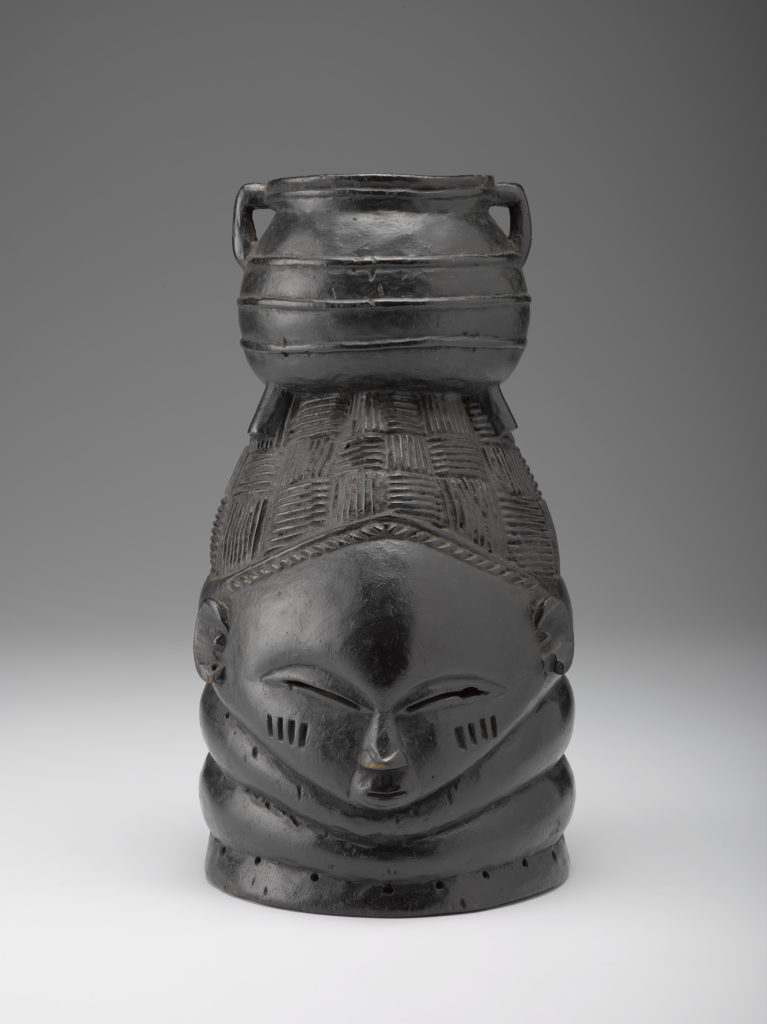 A black wooden figure of a human face with a pot balanced on top of the figure’s head.
