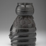 A black wooden figure of a human face with a pot balanced on top of the figure’s head. Side view.