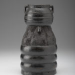 A black wooden figure of a human face with a pot balanced on top of the figure’s head. Reverse view.