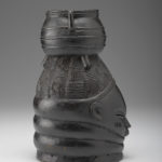 A black wooden figure of a human face with a pot balanced on top of the figure’s head. Side view.