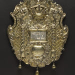 A gold shield featuring images of a crown and two columns, with three small bells hanging from the bottom edge.