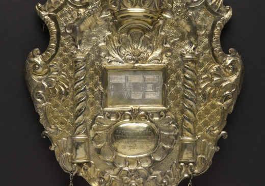 A gold shield featuring images of a crown and two columns, with three small bells hanging from the bottom edge.