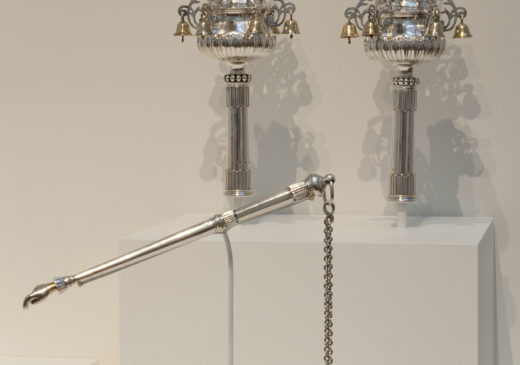 Two matching silver Torah finials, each topped with a gold crown and small gold bells that hang from intricate decorative elements. A matching pointer sits below, the tip is shaped like a hand pointing its finger and a chain extends from the end.