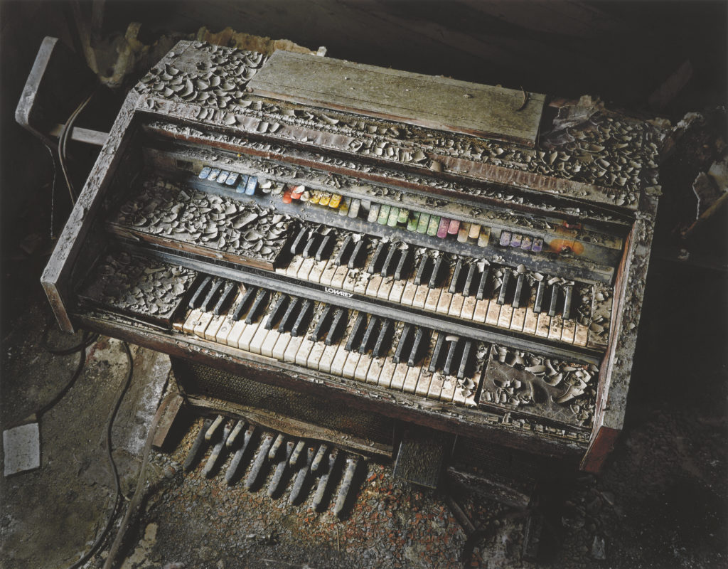 A color photograph of a water-damaged church organ covered in debris and surrounded by dark shadows.