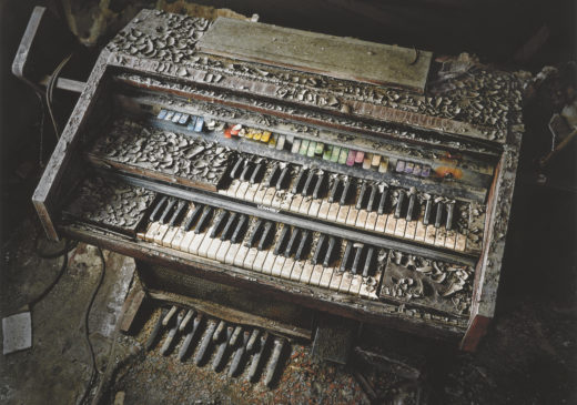 A color photograph of a water-damaged church organ covered in debris and surrounded by dark shadows.