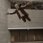 A sculpture of a man wearing a brown suit and a black necktie that is fluttering beside his head. The sculpture is suspended from the ceiling and appears to be falling from the sky.