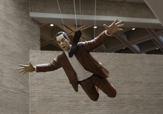 A sculpture of a man wearing a brown suit and a black necktie that is fluttering beside his head. The sculpture is suspended from the ceiling and appears to be falling from the sky.