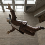 A sculpture of a man wearing a brown suit and a black necktie that is fluttering beside his head. The sculpture is suspended from the ceiling and appears to be falling from the sky. Alternate view.