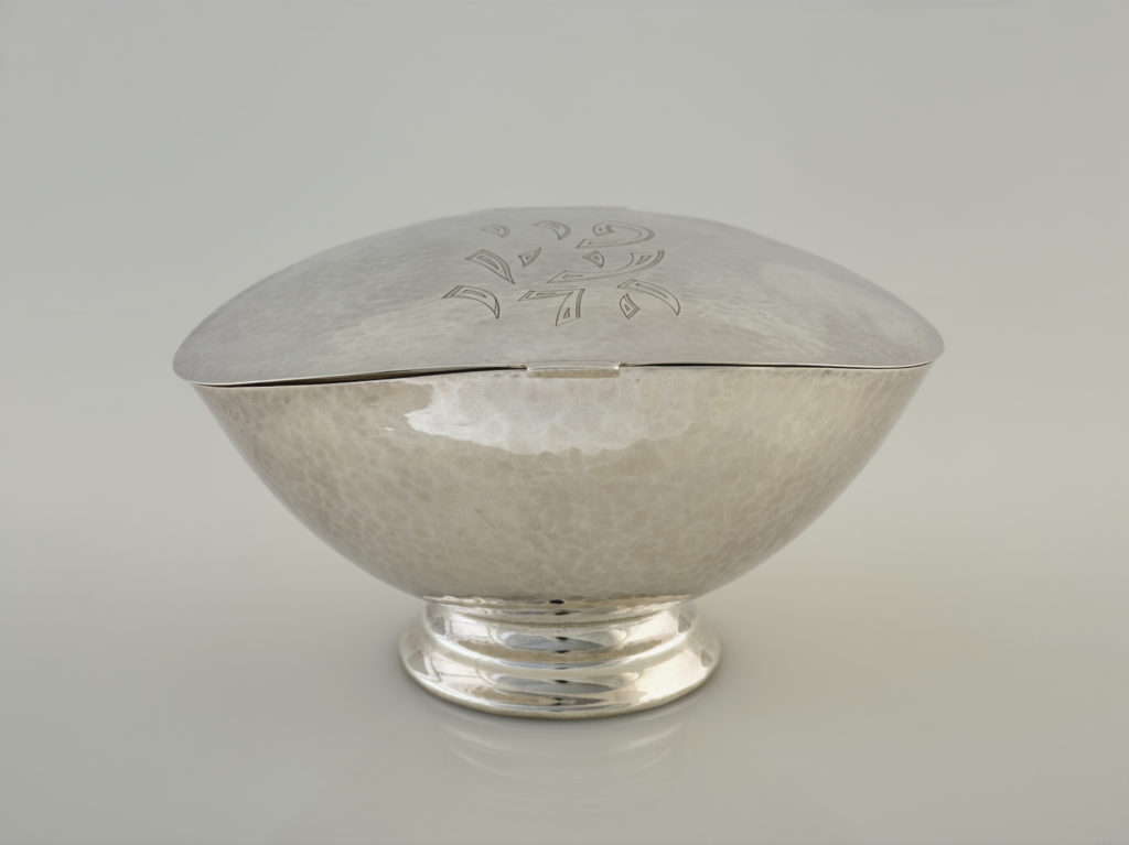 A silver, almond-shaped bowl with an attached lid and a round base. Hebrew text is engraved on top of the lid.