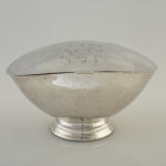 A silver, almond-shaped bowl with an attached lid and a round base. Hebrew text is engraved on top of the lid.