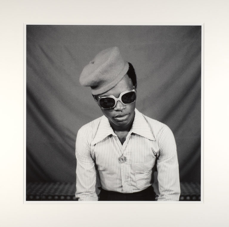 A black-and-white photograph of a man with a dark skin tone wearing a striped, button-down shirt with a wide collar, dark sunglasses, a hat, and a necklace with a round pendant.