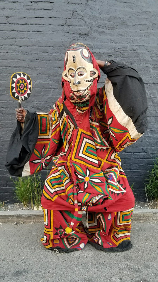 A red masquerade costume with a colorful pattern of flower shapes and nested rectangles, and wide sleeves with sections of solid white and black fabric. The ensemble includes a paddle-like fan with a sunburst design and a white face mask with black and red details. The features on the mask include wide eyes, bared teeth, and textured scars.