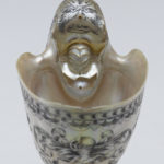 A decorative drinking cup made from an Indonesian nautilus shell. The body of the cup is decorated with a floral pattern. The cup’s stem is a carved oyster shell that is decorated with cupid figures and images of fruit. Detailed view.