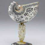 A decorative drinking cup made from an Indonesian nautilus shell. The body of the cup is decorated with a floral pattern. The cup’s stem is a carved oyster shell that is decorated with cupid figures and images of fruit. Alternate view.