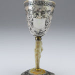 A decorative drinking cup made from an Indonesian nautilus shell. The body of the cup is decorated with a floral pattern. The cup’s stem is a carved oyster shell that is decorated with cupid figures and images of fruit. Alternate view.
