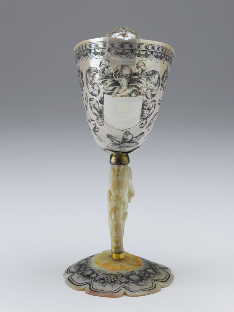 A decorative drinking cup made from an Indonesian nautilus shell. The body of the cup is decorated with a floral pattern. The cup’s stem is a carved oyster shell that is decorated with cupid figures and images of fruit.
