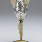 A decorative drinking cup made from an Indonesian nautilus shell. The body of the cup is decorated with a floral pattern. The cup’s stem is a carved oyster shell that is decorated with cupid figures and images of fruit. Alternate view.