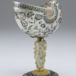 A decorative drinking cup made from an Indonesian nautilus shell. The body of the cup is decorated with a floral pattern. The cup’s stem is a carved oyster shell that is decorated with cupid figures and images of fruit. Alternate view.