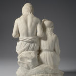 A white marble sculpture of a father and daughter sitting. The daughter sits slightly lower than the father while she works on a weaving project. Her father is carving an arrowhead. A slaughtered deer lies at their feet. View of reverse.