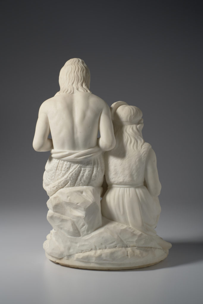 A white marble sculpture of a father and daughter sitting. The daughter sits slightly lower than the father while she works on a weaving project. Her father is carving an arrowhead. A slaughtered deer lies at their feet.