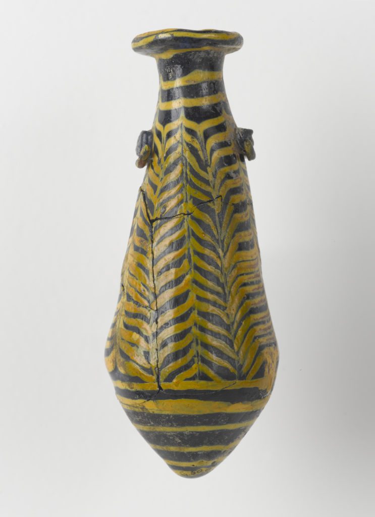 A small, dark-blue glass vessel with yellow designs. The vessel is long with a rounded bottom. It has two small rectangular handles on either side of the neck and a lipped opening.