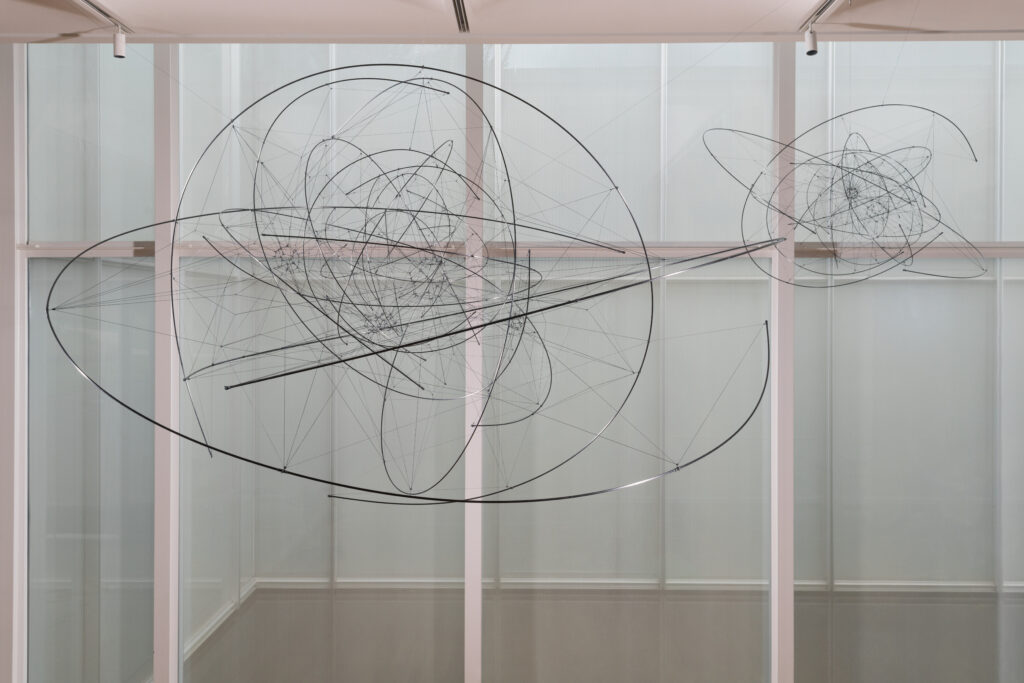 Two spherical sculptures, one larger and one smaller, hang suspended from a ceiling by wires in front of a wall of windows. The sculptures are made up of thin wires connecting suspended metal rings and half-rings.