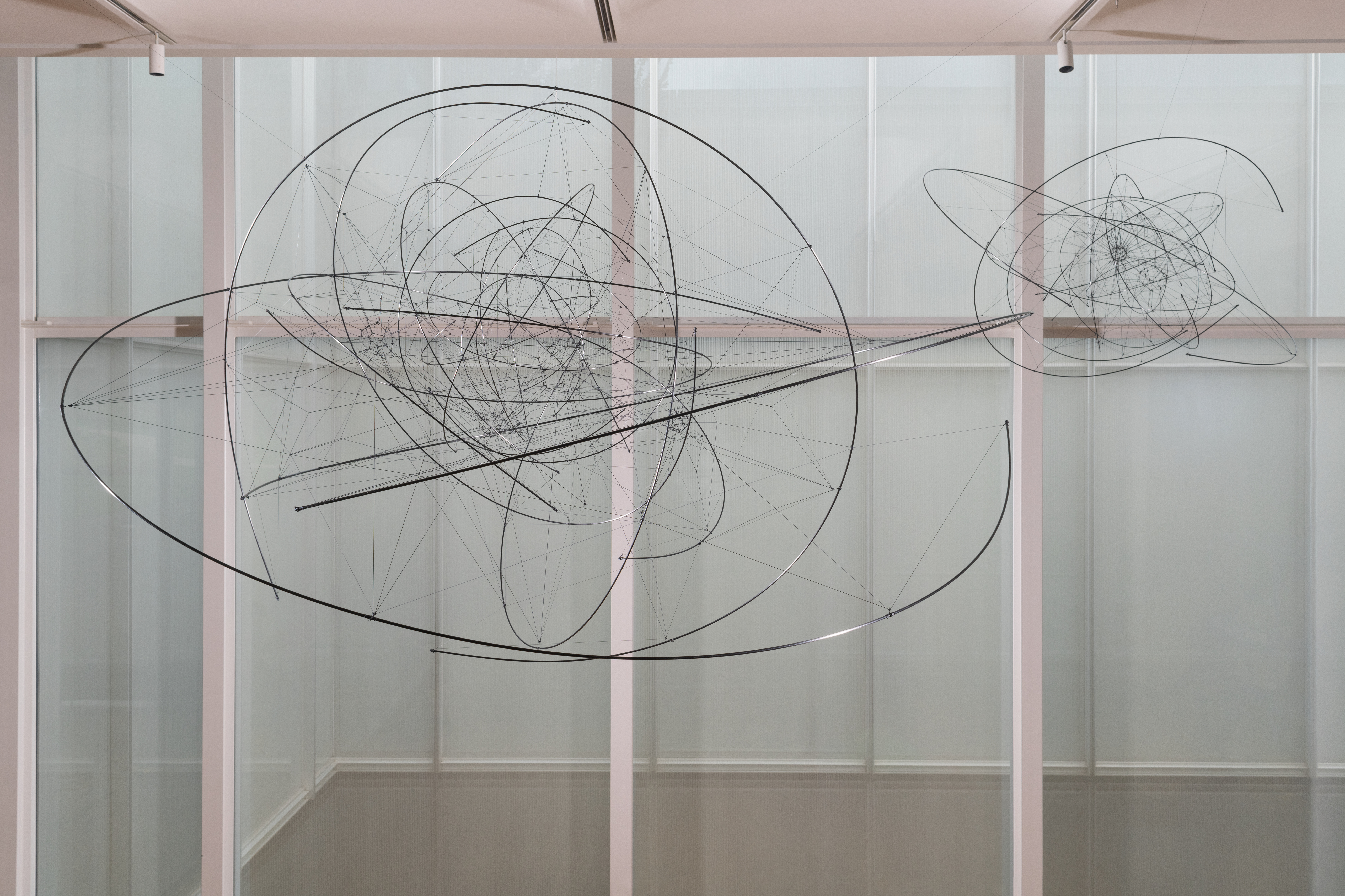 2022-09-08_SARACENO_Zonal_Harmonic_170,000_TR_2022_12_1 Two spherical sculptures, one larger and one smaller, hang suspended from a ceiling by wires in front of a wall of windows. The sculptures are made up of thin wires connecting suspended metal rings and half-rings.