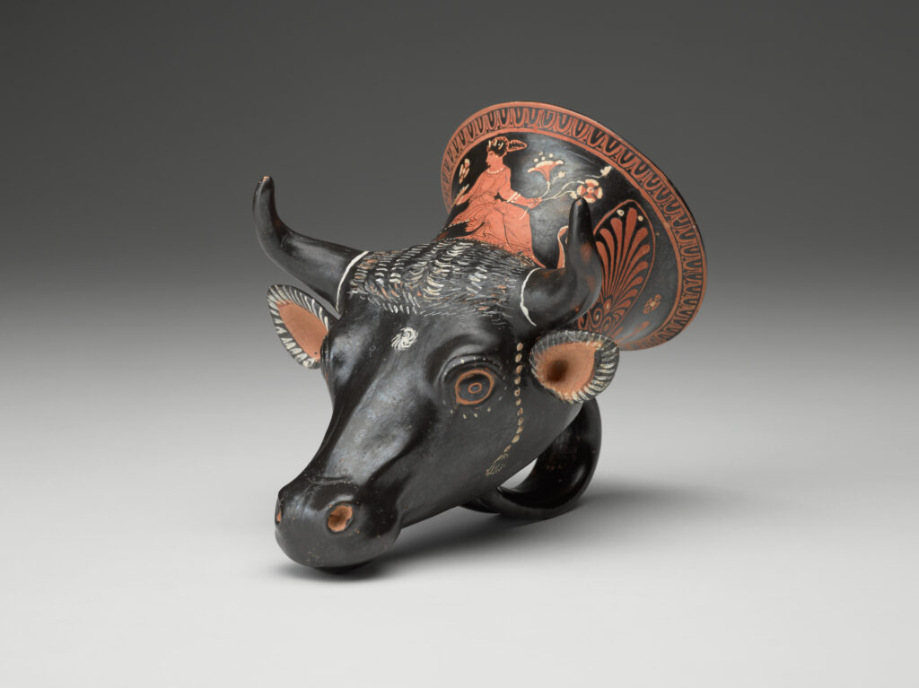 A black vase in the shape of a bull’s head, with a handle at the bottom and painted designs on the surface. View from side.