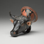 A black vase in the shape of a bull’s head, with a handle at the bottom and painted designs on the surface. View from side.