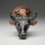 A black vase in the shape of a bull’s head, with a handle at the bottom and painted designs on the surface. View from front.