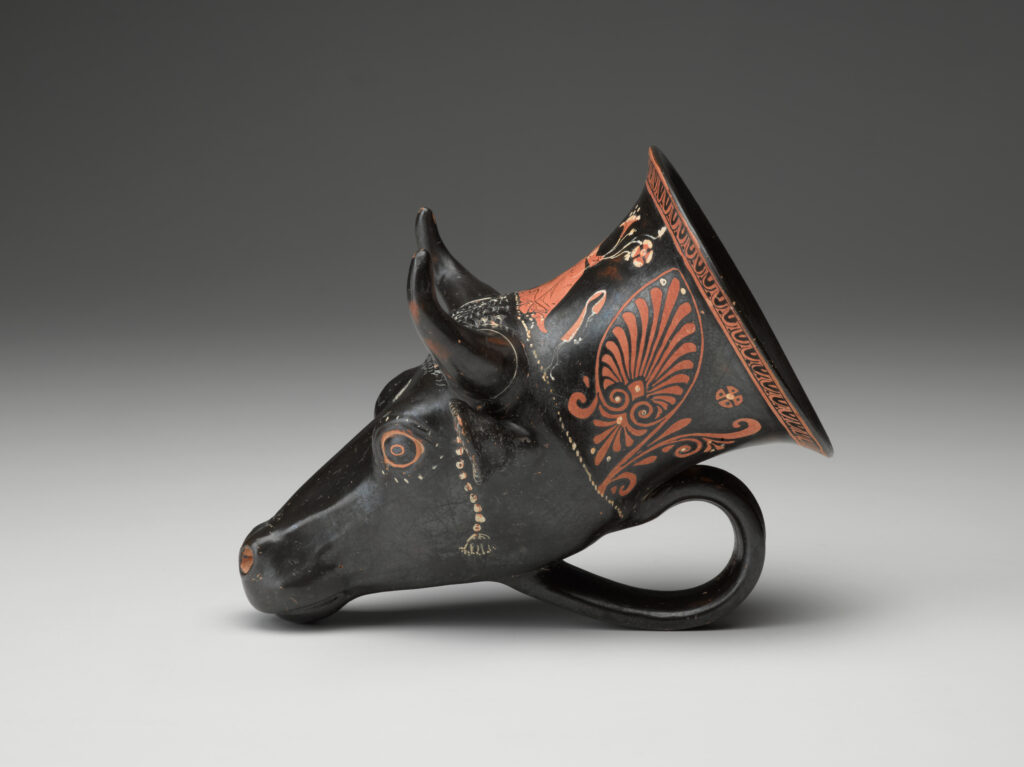 A black vase in the shape of a bull’s head, with a handle at the bottom and painted designs on the surface. View from side.