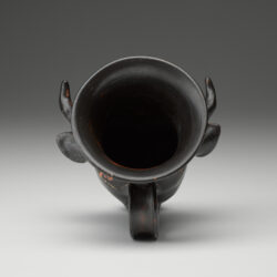 A black vase in the shape of a bull’s head, with a handle at the bottom and painted designs on the surface. View of opening.