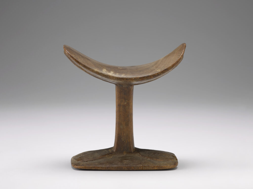 A wooden headrest that is similar in shape to a capital letter I. Both ends of the upper portion curl upward, to cradle a person’s head while they sleep.