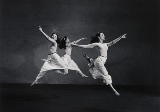A black-and-white photograph of three dancers wearing long white skirts. The three women are in mid-air, leaping away from each other with their arms extended upward.