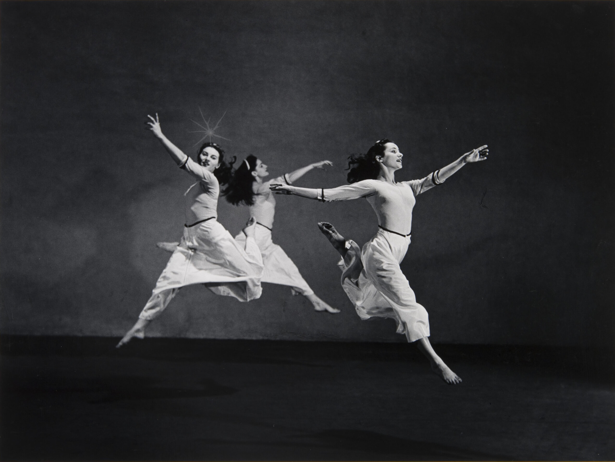 MORGAN, Martha Graham, American Document (Starburst), 2002_20_6 A black-and-white photograph of three dancers wearing long white skirts. The three women are in mid-air, leaping away from each other with their arms extended upward.