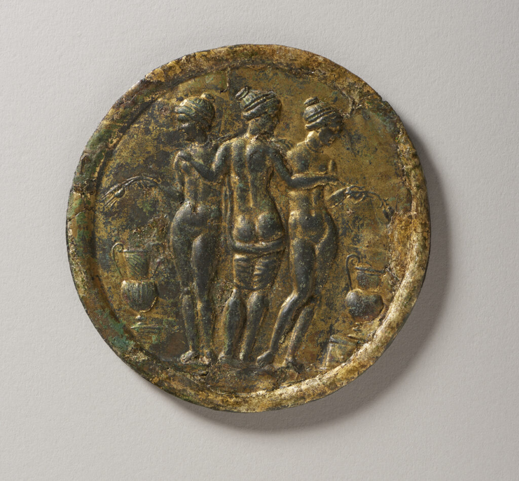 A round, gold-colored metal disk featuring three nude female figures who stand out against a flat background. The center figure is shown from the back and is embracing the other two figures, who are shown from the front. The figures are surrounded by images of bundled wheat and large water vessels with handles.