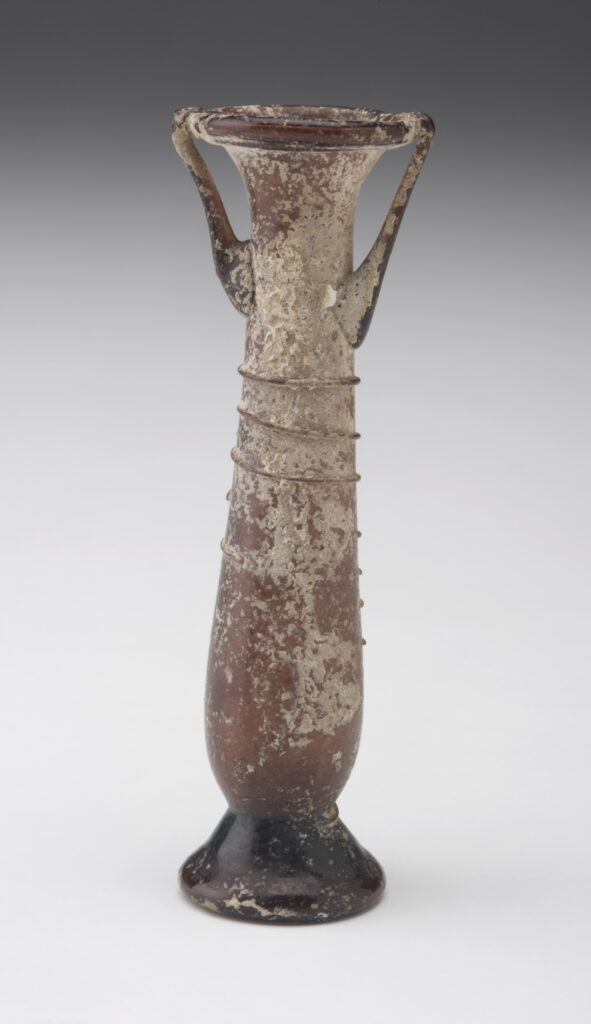 A tubular glass vessel with a long neck and two small handles. The vessel is a brownish-purple color and is decorated with a ribbon of glass that spirals around its neck. It has a flared base and a small opening.