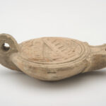A reddish-beige, ceramic object with a rounded body, spout, and small handle. Its concave top surface is decorated with a cityscape design. Side view.