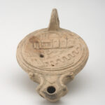 A reddish-beige, ceramic object with a rounded body, spout, and small handle. Its concave top surface is decorated with a cityscape design. View from above.