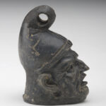 A small, black ceramic lantern in the shape of a man’s head wearing a cone-shaped hat. The face has deep wrinkles. There are holes in the eyes and under the ears, and the mouth is wide open. Side view